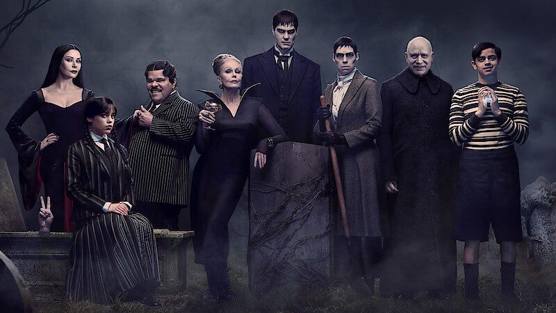 A group portrait of the Addams Family standing in a graveyard, featuring gothic attire and a moody atmospheric backdrop.