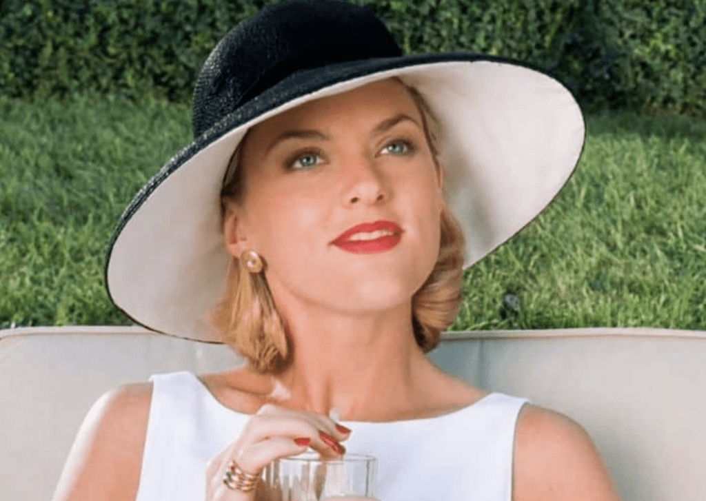A close-up of a woman wearing a large black and white hat, holding a drink. She has stylish makeup and is seated outdoors, exuding elegance and charm.