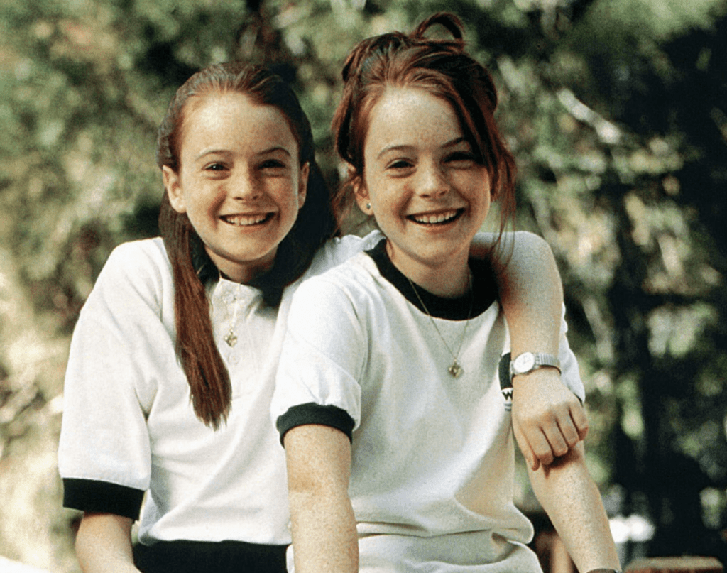 Two young girls sitting closely together, smiling joyfully, dressed in matching black and white outfits, surrounded by a natural setting.