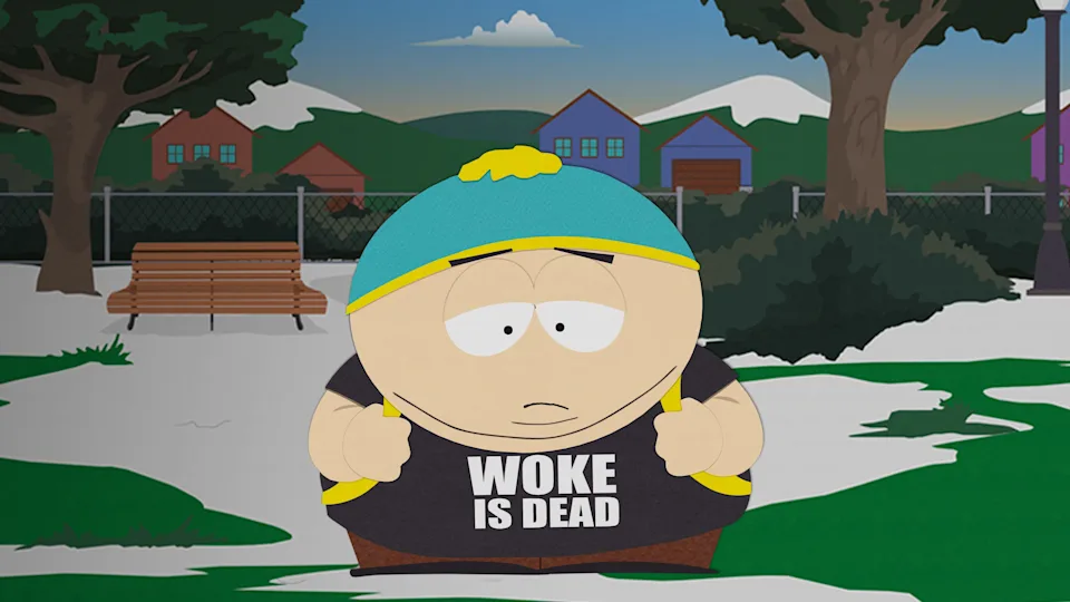 Cartman from South Park stands in a snowy park wearing a black shirt that reads 'WOKE IS DEAD'. The background features a bench and colorful houses.